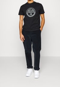 Camiseta negra de algodón con un logo gráfico circular gris en el pecho, combinada con pantalones de pana oscuros y zapatillas blancas.