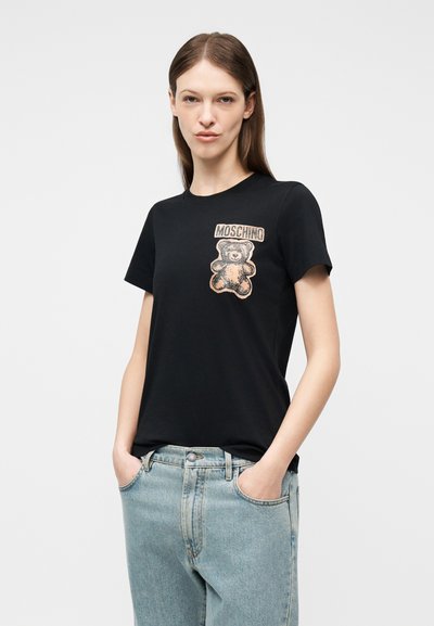 Νέα γυναίκα που φοράει μαύρο T-shirt Moschino με γραφικό αρκουδάκι και ανοιχτόχρωμο μπλε τζιν, με τα χέρια στις τσέπες, μπροστά σε απλό φόντο.