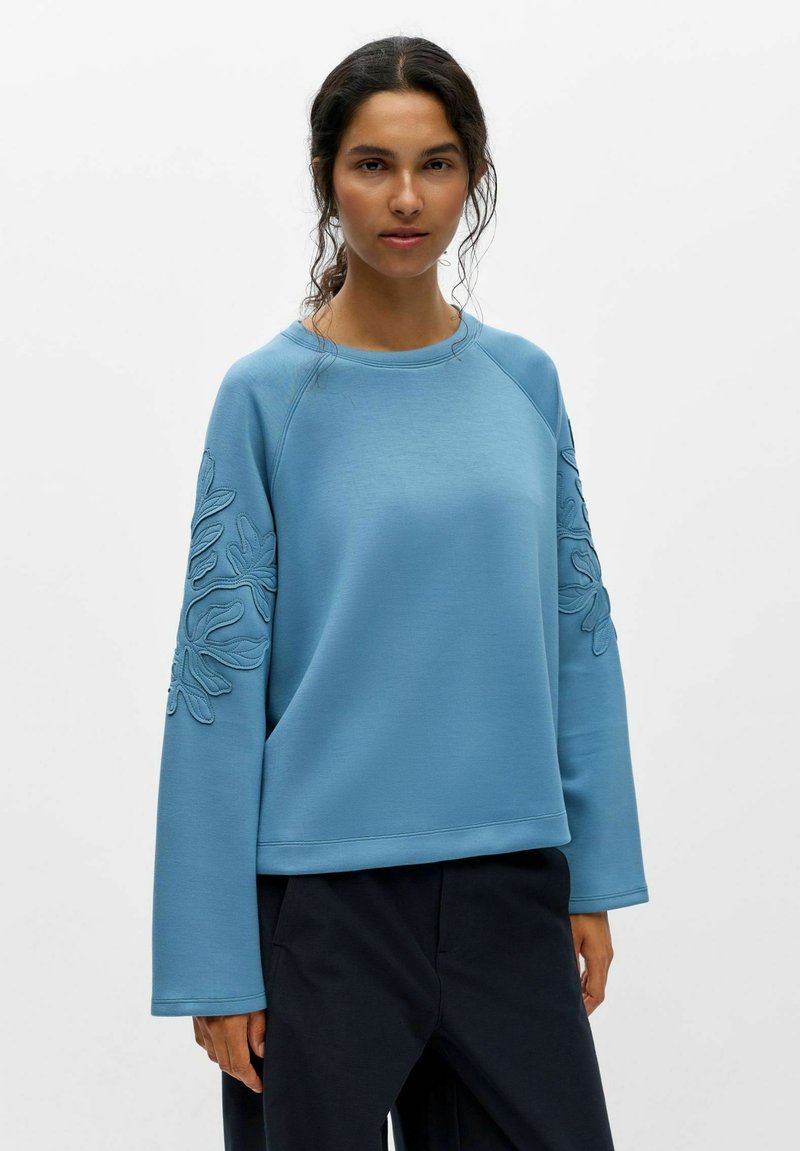 Femme aux cheveux foncés porte un sweat-shirt bleu clair ample avec broderie de feuilles sur les manches et un pantalon noir sur fond blanc uni.