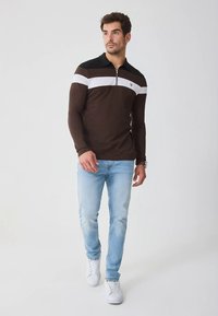 Bruine lange mouwen polo met een wit-zwarte streepdessin, kraag en halve rits. Gecombineerd met lichtblauwe jeans en witte sneakers.