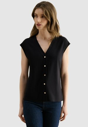 Mujer con cabello ondulado hasta los hombros que lleva una blusa negra de manga corta con botones y vaqueros azul oscuro, mirando ligeramente hacia un lado.
