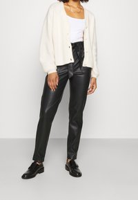 Cardigan crémeux en peluche avec des boutons, associé à un pantalon en cuir synthétique noir et des chaussures noires. Design simple et décontracté.