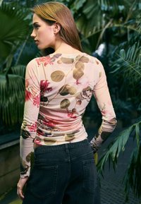 Langärmliges, florales Oberteil in zartem Rosa mit grünen Blättern, featuring eine figurbetonte Passform und gerafften Rücken. Kombiniert mit dunklen Jeans.