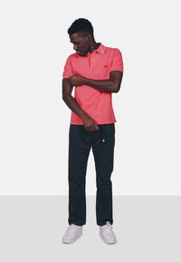 Rotes Poloshirt mit Kragen, kurzen Ärmeln und kleinem Logo, kombiniert mit schwarzen Hosen und hellen Turnschuhen. Der Stoff scheint glatt zu sein.