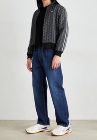 Czarna i szara kurtka w wzory z prążkowanymi mankietami i kołnierzem, w połączeniu z niebieskimi dżinsami i białymi butami sportowymi z niebieskimi akcentami.