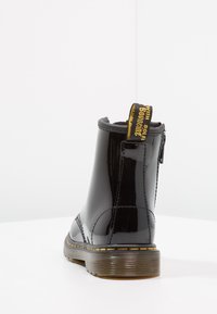 Botas de tobillo de charol negro con una lengüeta amarilla con marca, detalle de cremallera y suela de goma texturizada. Acabado suave y puntera redondeada.