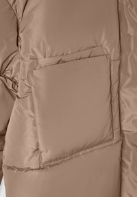 Giubbotto imbottito di un marrone chiaro con una texture liscia, caratterizzato da una tasca quadrata sul davanti e dettagli cuciti lungo le cuciture.