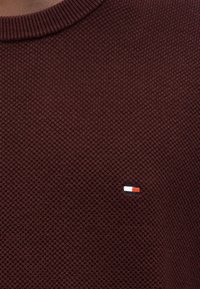 Μπορντό πλεκτό πουλόβερ με ανάγλυφο σχέδιο και στρογγυλή λαιμόκοψη. Διαθέτει μικρό λογότυπο σε κόκκινο, λευκό και μπλε στο στήθος.