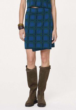 Femme portant une jupe à carreaux bleue et verte, des bottes hautes en daim marron et deux bracelets au poignet gauche.