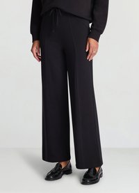 Pantalon noir à jambes larges avec taille à cordon, présentant un tissu texturé et des détails de coutures verticales. Associé à des mocassins noirs.