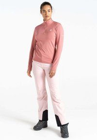 Langärmliger, halber Reißverschluss-Pullover aus hellrosa Stoff. Kombiniert mit hellrosa Hosen mit schwarzen Akzenten und robusten schwarzen Wanderschuhen.