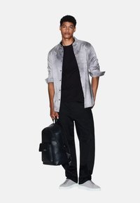 Camicia grigia a bottoni, t-shirt nera, pantaloni neri, scarpe grigie slip-on, zaino nero con tasca frontale, in piedi su uno sfondo semplice.