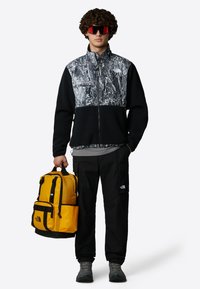 Czarna polarowa kurtka z abstrakcyjnym szarym wzorem, czarne spodnie cargo, szara koszula, czerwone okulary przeciwsłoneczne i żółczny plecak z czarnymi akcentami.