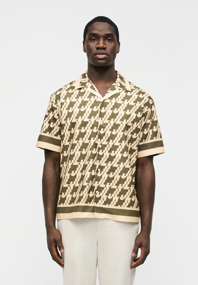 Man wearing a beige and olive green patterned short-sleeve button-up shirt paired with light-colored pants, standing against a plain background.