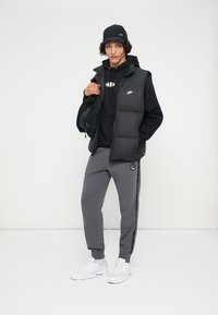 Colete puffer preto sobre um hoodie preto, calças de moletom cinzentas com riscas laterais, e sapatilhas brancas. Acessórios incluem um boné preto e uma mala.