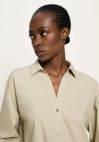 Woman with braided hair in a beige buttoned shirt looking sideways against a plain light background.