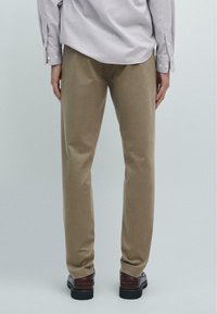 Beige bukser med en glat tekstur, lige ben design og subtil glans; parret med mørkebrune sko. Synlig ryg af en stribet skjorte.