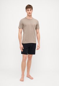 T-shirt en coton beige avec logo rouge "HUGO" sur la poitrine, assortie à un short noir. Le modèle se tient pieds nus devant un fond blanc.