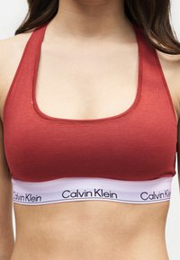 Femme portant un soutien-gorge de sport Calvin Klein couleur rouille avec une bande élastique blanche à logo, montrée des épaules jusqu'au ventre.