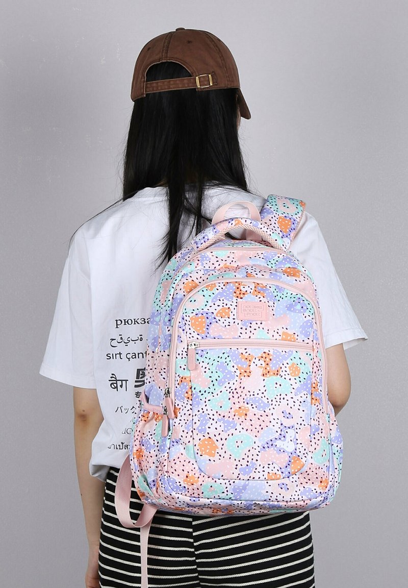 Personne portant un sac à dos coloré à motif pastel, un T-shirt blanc avec texte, une casquette marron et un pantalon rayé noir et blanc, vue de dos.