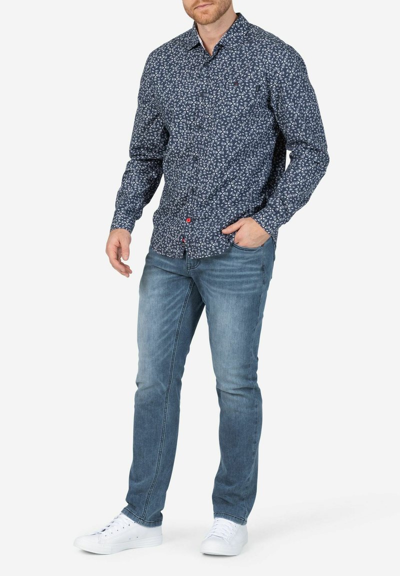 Marineblauw overhemd met bloemenpatronen, lange mouwen en rode knoopaccenten, gecombineerd met lichtblauwe jeans en witte sneakers.