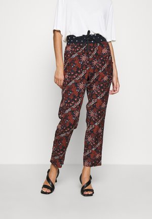 Kvinde iført bukser med højt talje og mønster i sort, rust og hvidt blomsterdesign, kombineret med sorte hælede sandaler og en hvid top.