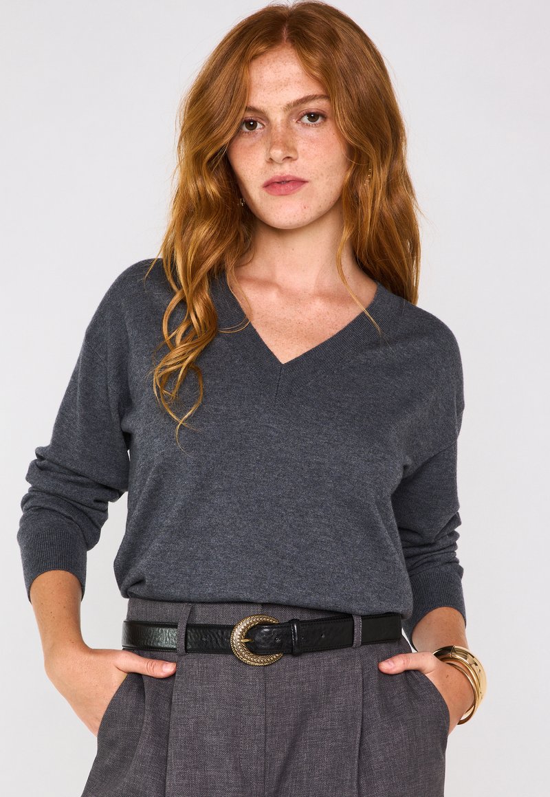 Un pull en V gris fabriqué dans un matériau doux, avec des manches longues, associé à un pantalon à carreaux taille haute et une ceinture noire avec une boucle en or.