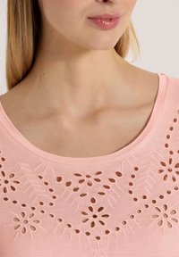 Hellrosa Baumwolloberteil mit floralen Ausschnitten, weitem Ausschnitt und glatter Textur, verziert mit dekorativen Nähten.