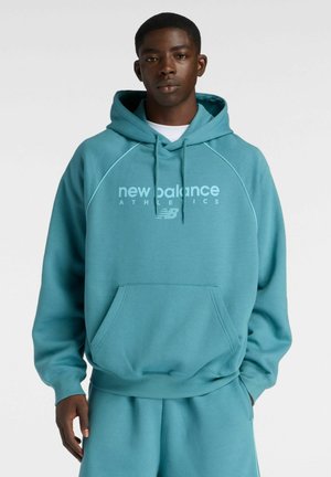 Junger Mann trägt einen türkisen New Balance Hoodie und passende Jogginghose, steht vor einfarbigem Hintergrund mit neutralem Gesichtsausdruck.