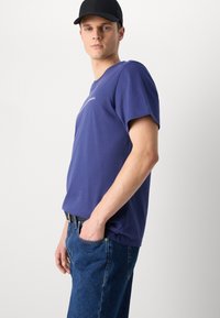 Navy blue cotton t-shirt with short sleeves, featuring a subtle logo on the chest, paired with blue denim jeans and a black cap.