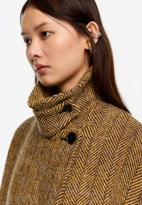 Abrigo de espiga marrón y amarillo con un alto cuello, dos botones negros y una textura suave. La modelo muestra el diseño del abrigo.