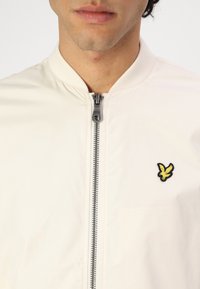 Lys creme bomberjakke med frontlynlås, rund krave og et gult og sort broderet logo på venstre bryst.