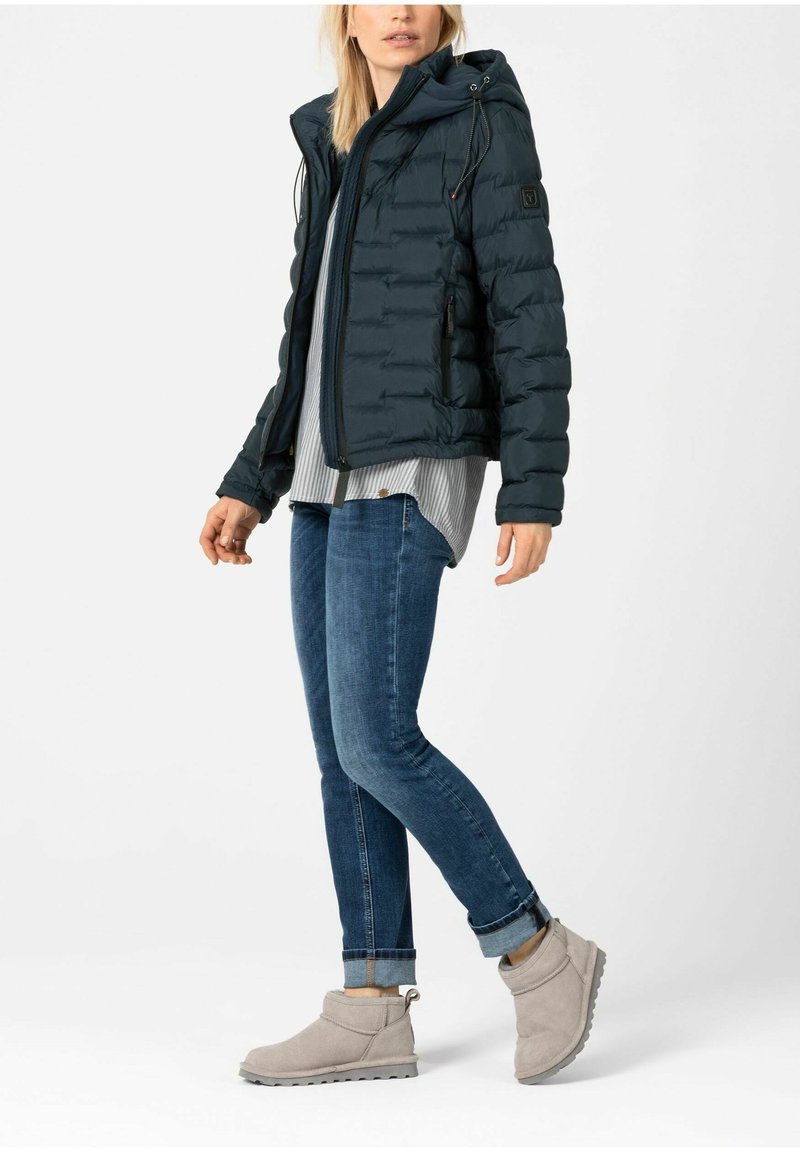 Dunkelgrüne Daunenjacke mit Kapuze, Reißverschluss vorne und gerippten Bündchen, kombiniert mit einem grau-weiß gestreiften Shirt und blauen Jeans. Graue Stiefeletten.