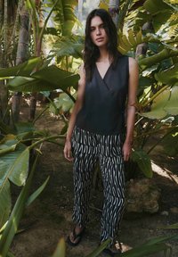 Donna con lunghi capelli scuri che indossa un top nero senza maniche e pantaloni a righe bianco-neri, in piedi tra grandi piante tropicali all'aperto.