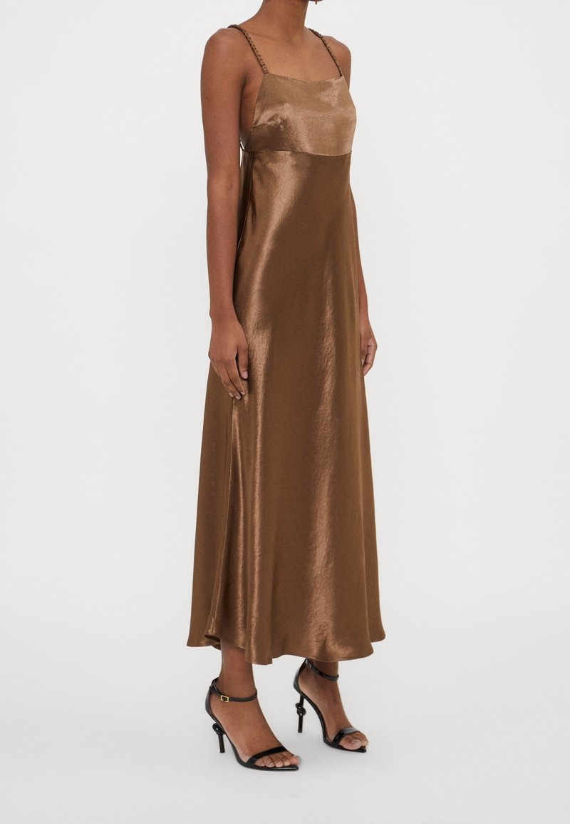 Femme portant une robe midi marron brillante sans manches avec de fines bretelles et des sandales noires à talons hauts, debout devant un fond uni.
