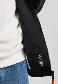 Black jacket with zippered closure, featuring windproof and water-repellent labels on the interior. Soft texture, paired with a white sweater.