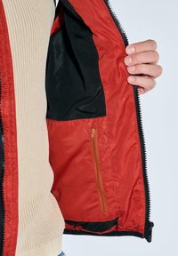 Rote Jacke mit schwarzem Innenfutter, ausgestattet mit einer Reißverschlusstasche, darunter sichtbar ein beiges, geripptes Hemd. Reißverschluss an der Vorderseite.