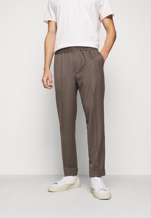 Man draagt bruine taps toelopende broek met omgeslagen zoom, wit T-shirt met ronde hals en witte lage sneakers, staand tegen een effen achtergrond.