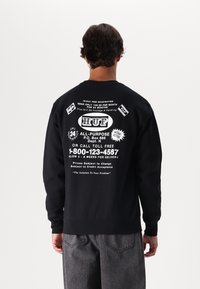 Felpa nera con testo e loghi grafici bianchi sul retro. Maniche lunghe con polsini e orlo a coste. Design semplice e vestibilità comoda.
