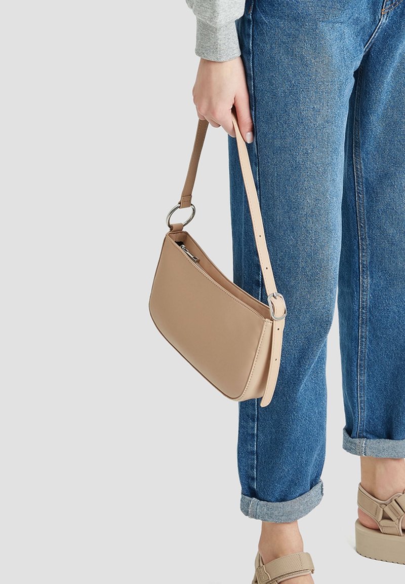 Personne portant un jean bleu retroussé, des sandales à plateforme beiges et tenant un petit sac à main en cuir beige avec un anneau argenté.