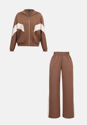 Ensemble de survêtement marron en deux pièces avec des rayures diagonales blanches sur les manches de la veste et des panneaux latéraux blancs sur le pantalon à jambes larges.