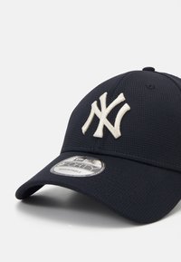 Cappello regolabile blu navy in tessuto testurizzato, con logo ricamato bianco dei New York Yankees. Include un adesivo circolare sulla visiera.