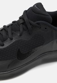 Chaussure de sport noire avec tige en mesh et semelle en caoutchouc lisse. Comprend des accents texturés et un logo Nike noir bien en vue.
