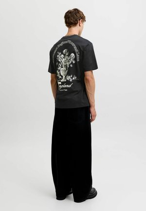 Ung mand står med ryggen til iført en sort T-shirt med hvidt blomstervaseprint og tekst, sat sammen med løse sorte bukser og sorte sko.