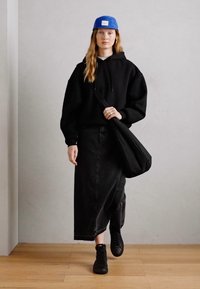 Haut noir à capuche, longue jupe en denim noir, baskets noires et casquette bleue. Porte un sac à bandoulière noir. Fond neutre et sol en bois.