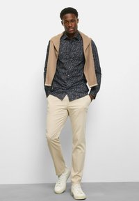 Dunkles, geblümtes Hemd mit Knöpfen, beigefarbener Pullover locker über die Schultern gelegt, hellbeige Hose und weiße Sneaker. Glatte Textur und tailliertes Design.