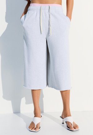 Személy világosszürke, bőszárú, rövidített melegítőnadrágot visel oldalzsebekkel és fehér sarokszandált, fehér felületen áll.