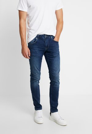 Slanke denim jeans in een donkere blauwe wassing, met vijf zakken en subtiele vervaagde details, gecombineerd met een eenvoudig wit t-shirt en witte sneakers.