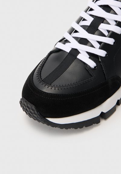 Zapatilla de cuero negro y ante con cordones blancos, que presenta una suela texturizada y costuras a lo largo de la puntera y los laterales.