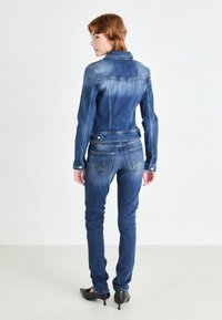 Veste en denim et jeans ajustés bleu moyen, avec des décolorations, poches arrière et accessoires en métal argenté. Porté avec des chaussures à talons noires.